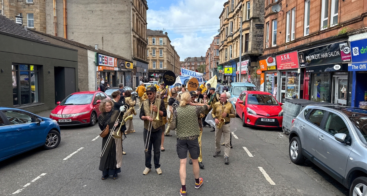 Parade participants dancing and carrying banners through the streets of Govanhill